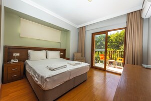 Habitación de hotel con una cama grande, una mesita de noche de madera, una silla y un balcón con vistas a zonas verdes.
