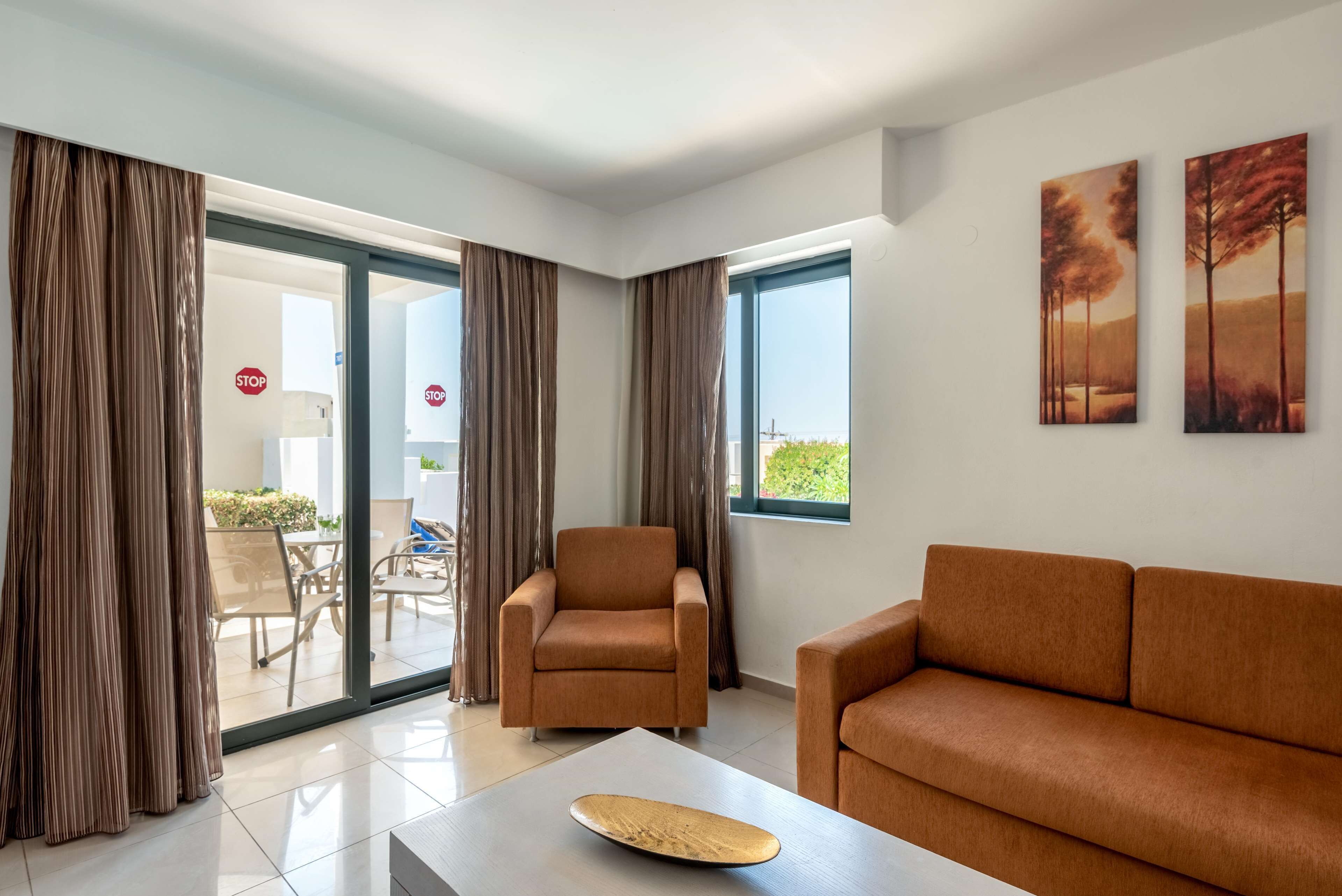 Un salon moderne avec un canapé, un fauteuil et une table sur laquelle se trouve un objet en bois. On y voit des rideaux, une vue à travers une porte vitrée et deux tableaux accrochés au mur.
