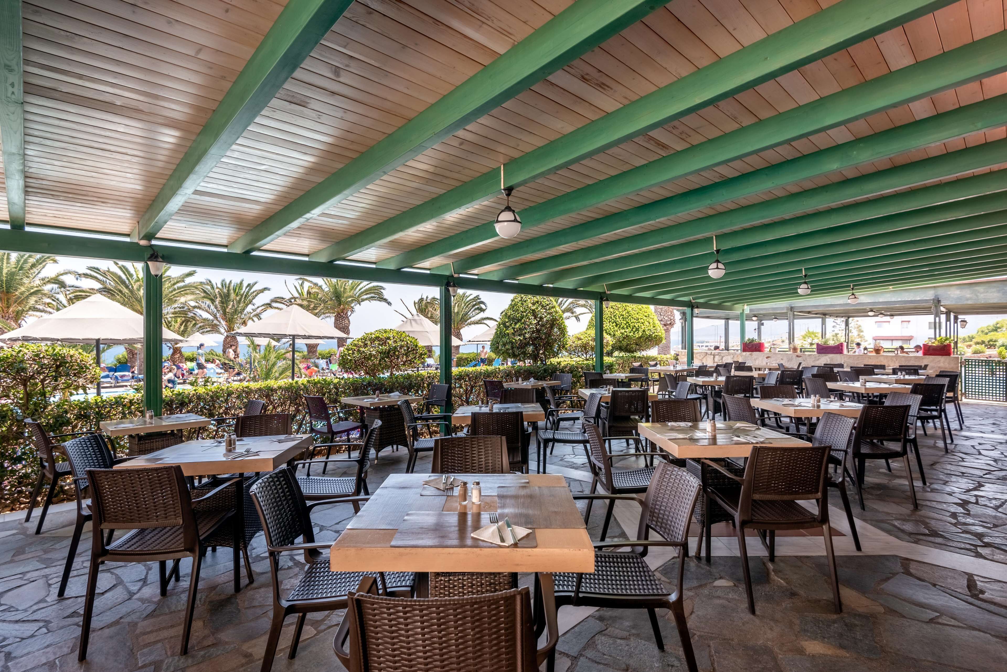 Un espace extérieur aménagé en terrasse, avec plusieurs tables et chaises, un plafond en bois et une vue sur des palmiers et autres plantes verdoyantes en arrière-plan.