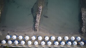 An aerial view of a beach with a stone pier extending into the water, surrounded by rows of blue chairs and white umbrellas. - Roda Beach Resort & Spa All-inclusive (Corfu)