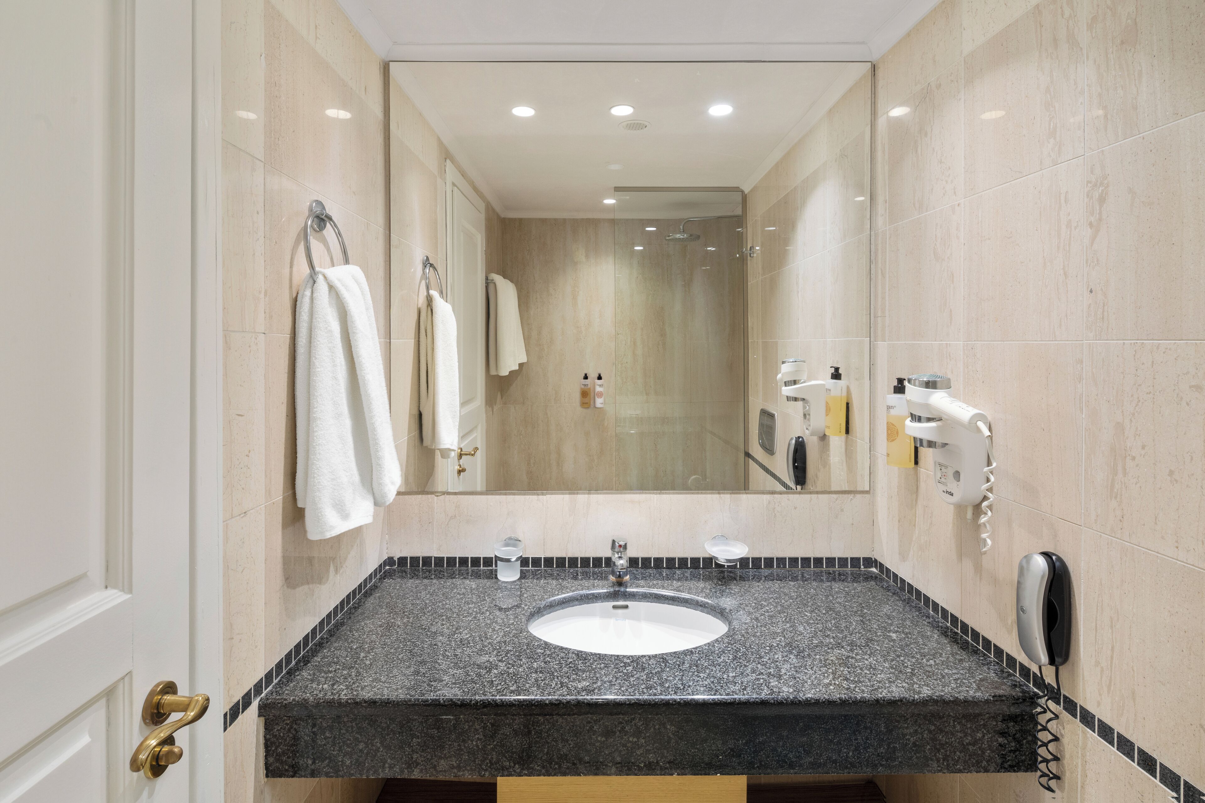Ein modernes Badezimmer mit einer Granittheke, einem Waschbecken und einem Spiegel. Handtücher hängen an der Wand und ein Föhn ist dort angebracht.