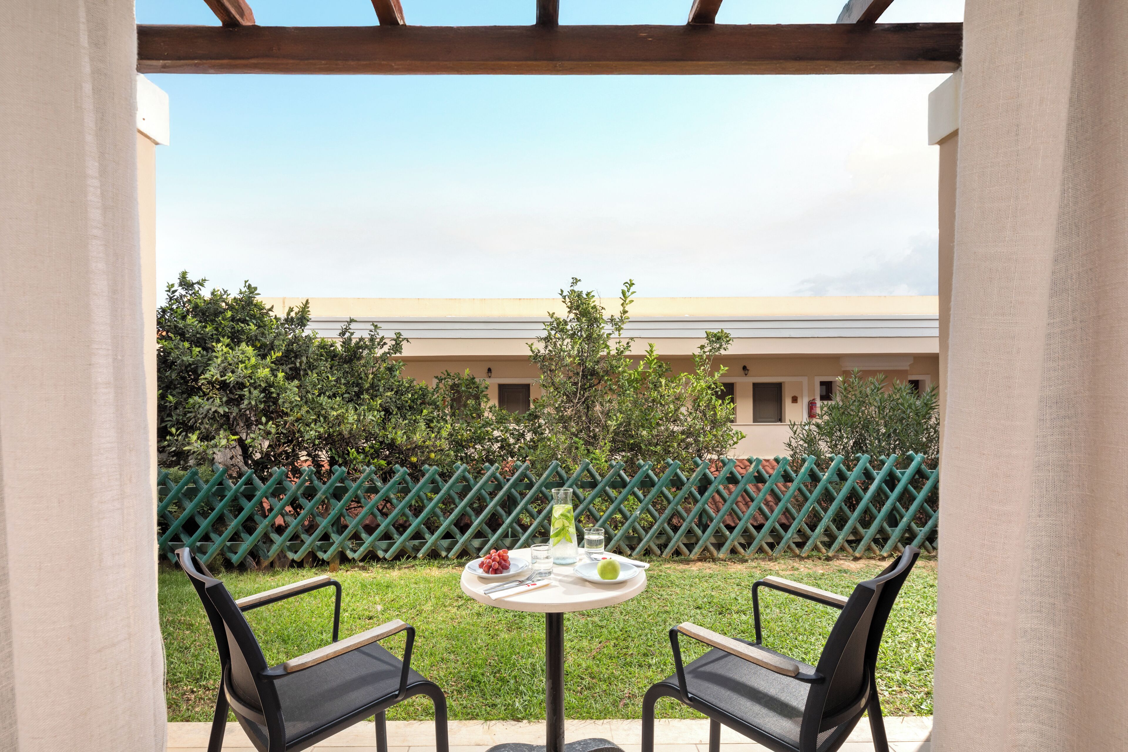 Un patio al aire libre con dos sillas y una mesa, una cerca verde y un edificio al fondo.