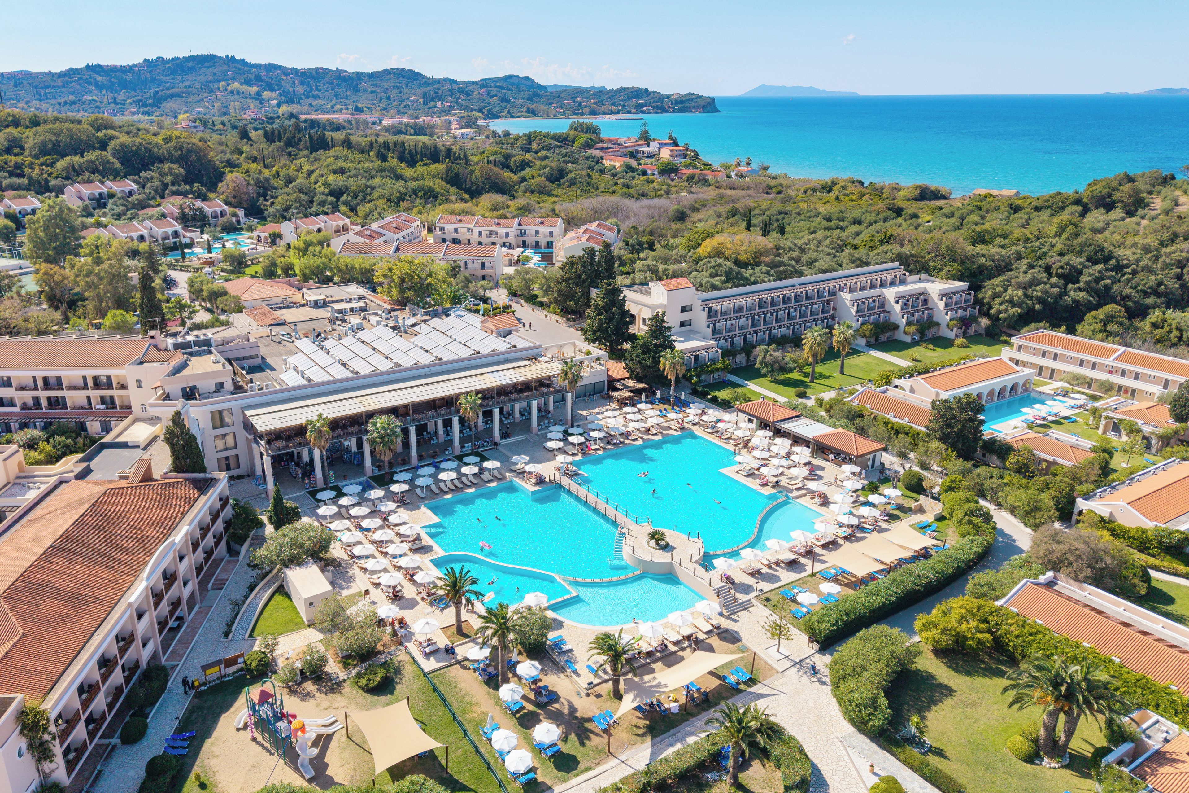 A resort with a central swimming pool surrounded by sun loungers and umbrellas, a children's play area, and a view of the sea.