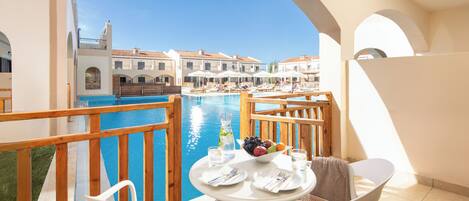 A balcony with a table set for two, overlooking a swimming pool and a building with a tiled roof.