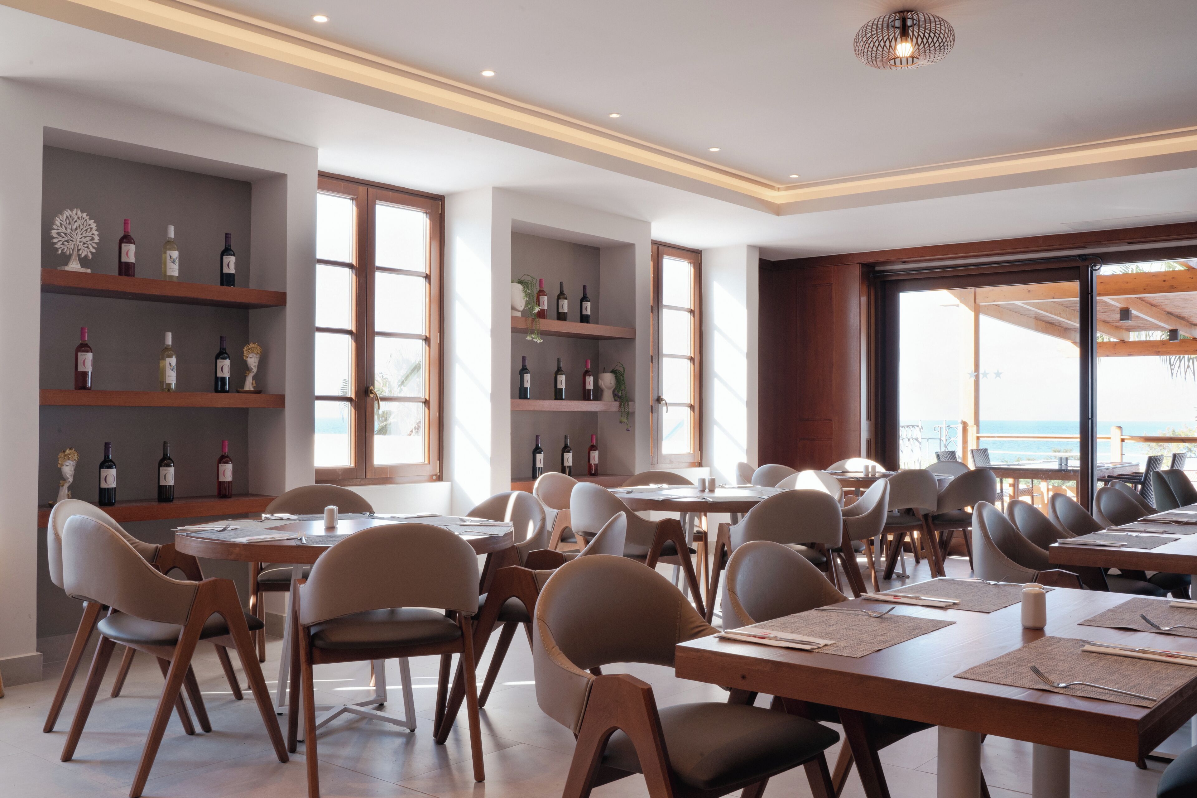 Un restaurante con varias mesas y sillas, estanterías llenas de botellas de vino y grandes ventanales con vista al mar.