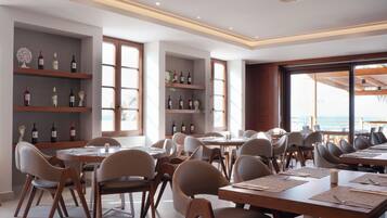 Un restaurante con varias mesas y sillas, estanterías llenas de botellas de vino y grandes ventanales con vista al mar.