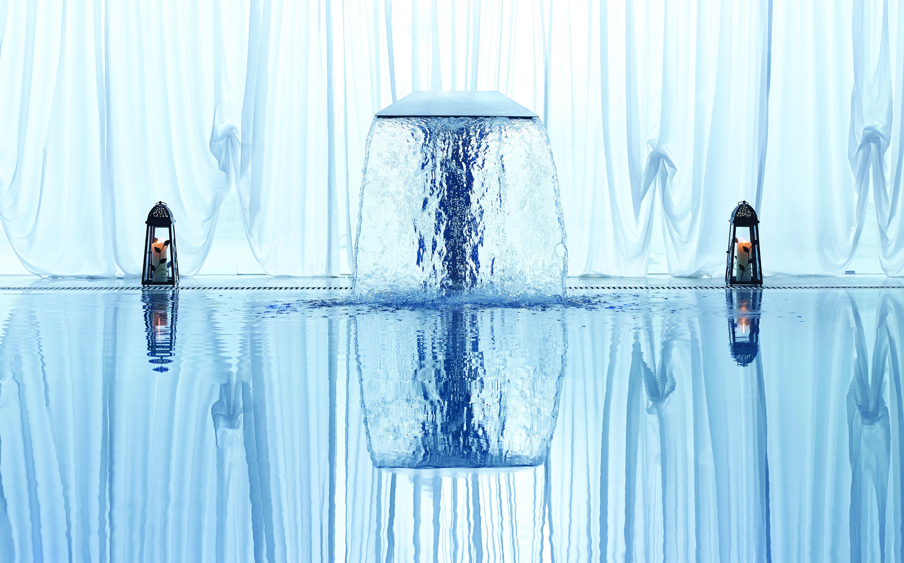 Una piscina interior tranquila con un elemento acuático central, rodeada de cortinas blancas y translúcidas.