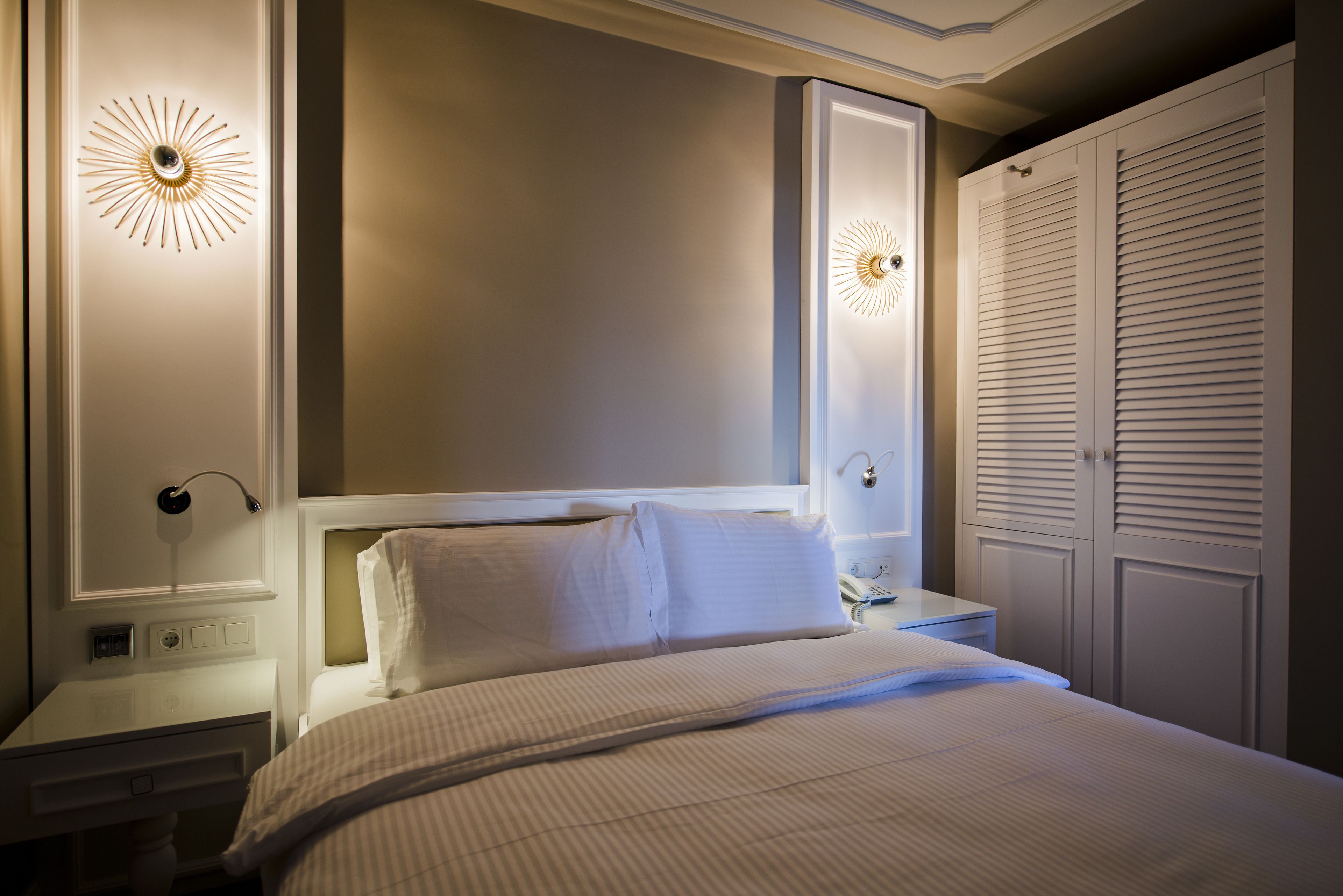 A neatly made bed with white linens, a bedside table with a lamp, and a wall-mounted light fixture.