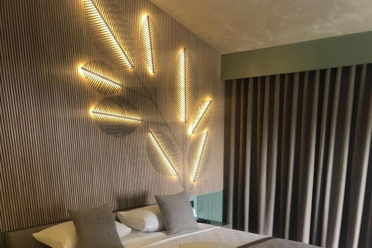 A modern bedroom with a large bed, a headboard with illuminated patterns, and a view of the outdoors through curtains.