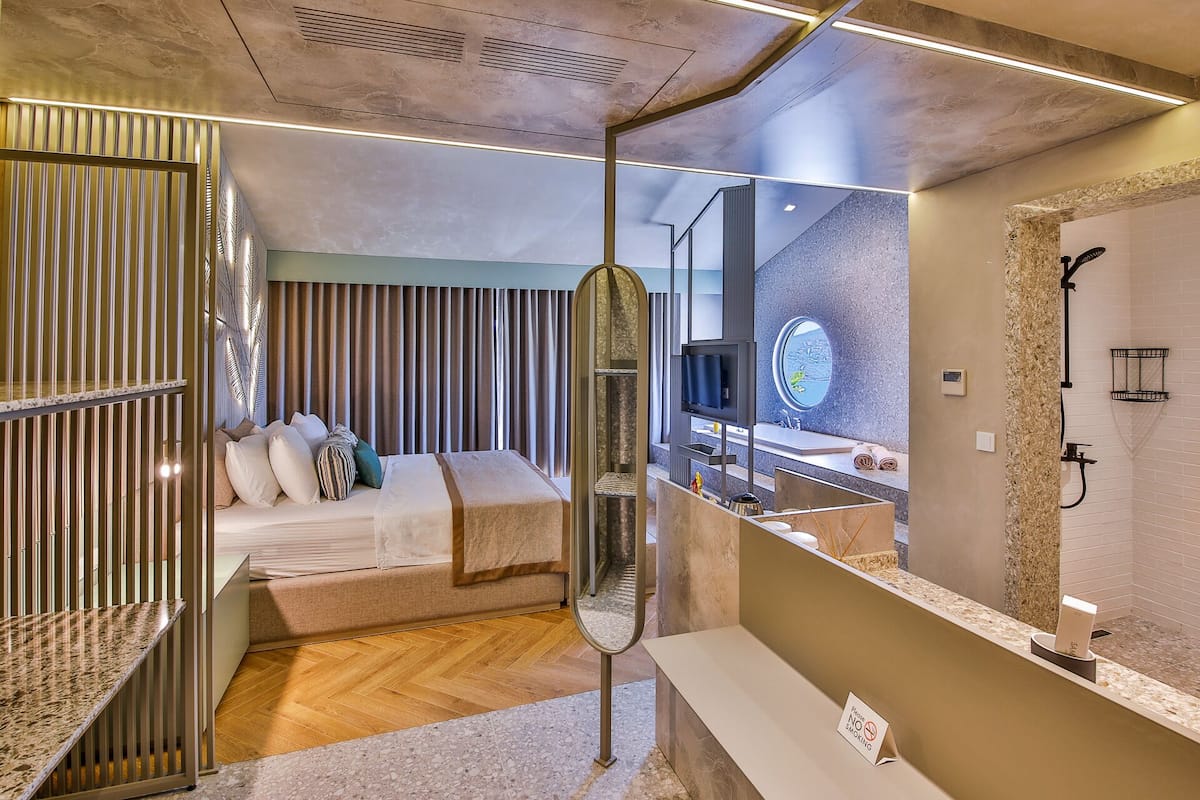 A modern hotel room with a bed, a TV, a bathroom with a shower, and a mirror.