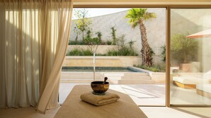 Un cuarto de spa con una camilla de masaje, un cuenco de madera y vistas a un jardín ajardinado a través de amplias puertas de cristal.