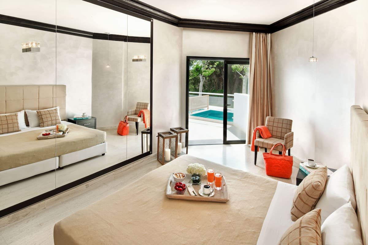 A modern hotel room with a bed, a sofa, a dining table with breakfast items, a chair, and a view of a swimming pool.