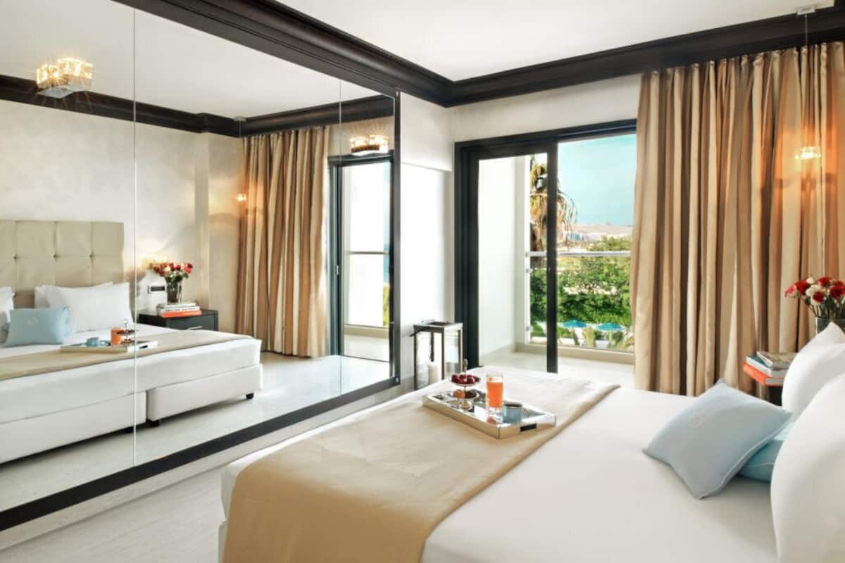 A modern hotel room with a large bed, a sofa, a dining table with breakfast items, a large window with curtains, and a balcony with a view.