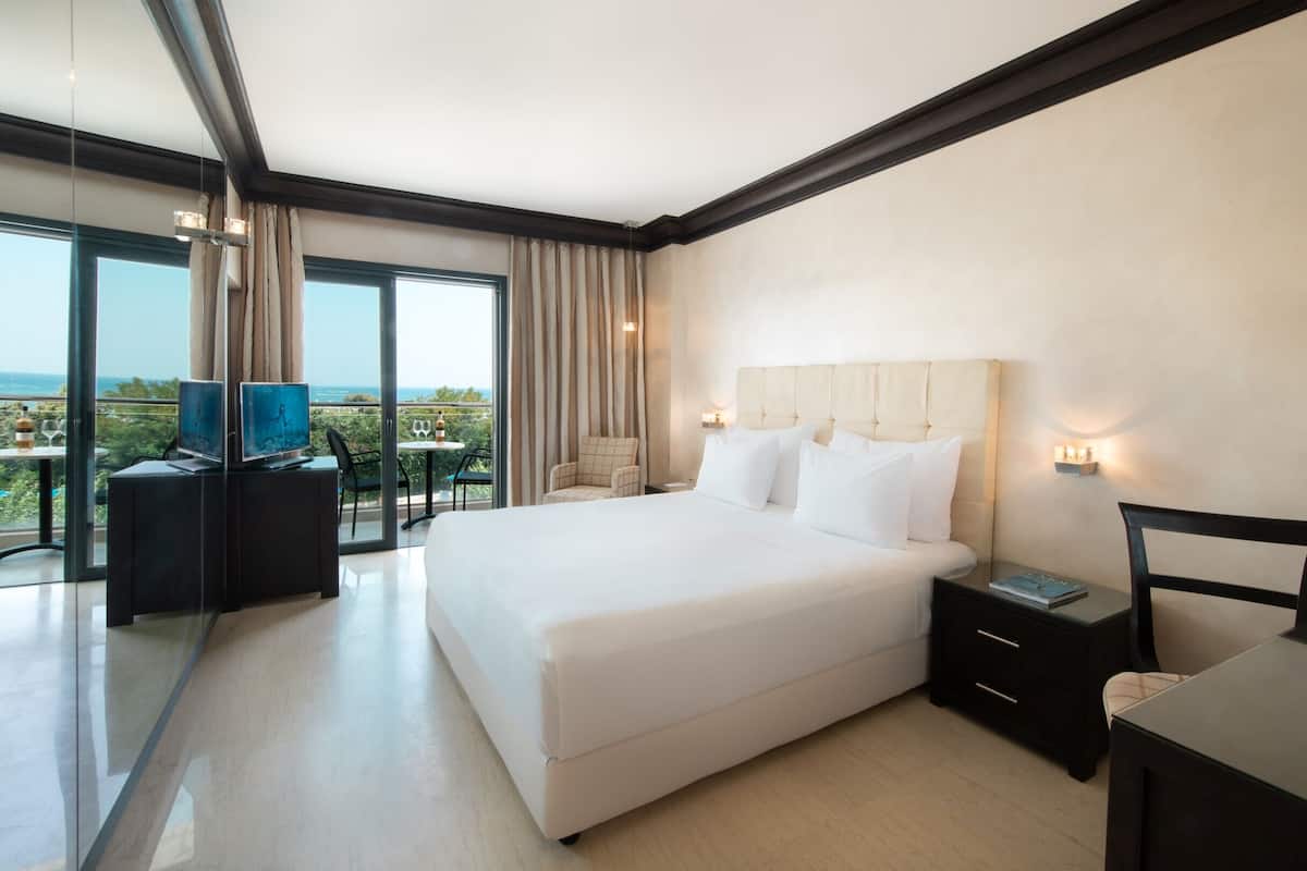 A modern hotel room with a large bed, a desk, and a chair. There is a balcony with a view.