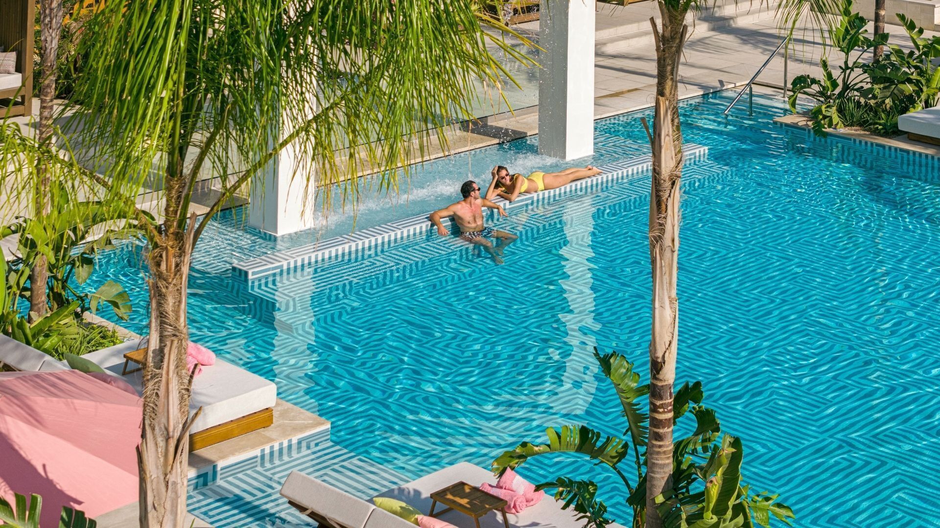 En swimmingpool med krystalklart, blåt vand, omgivet af palmer og liggestole.
