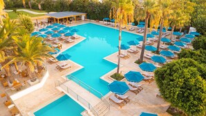 Una amplia piscina rectangular con varios niveles de solárium, rodeada de palmeras y sombrillas azules.