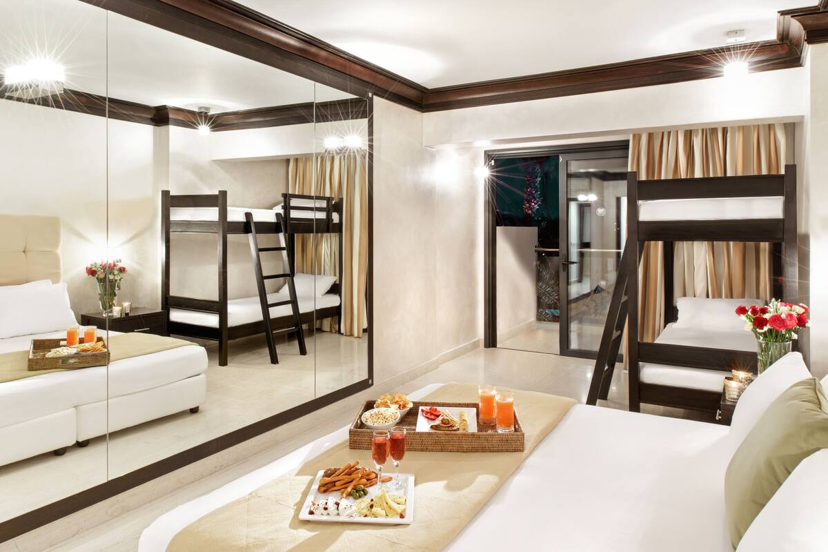 A hotel room with a sofa, a dining table with snacks and drinks, a bunk bed, and a large mirror reflecting the room.