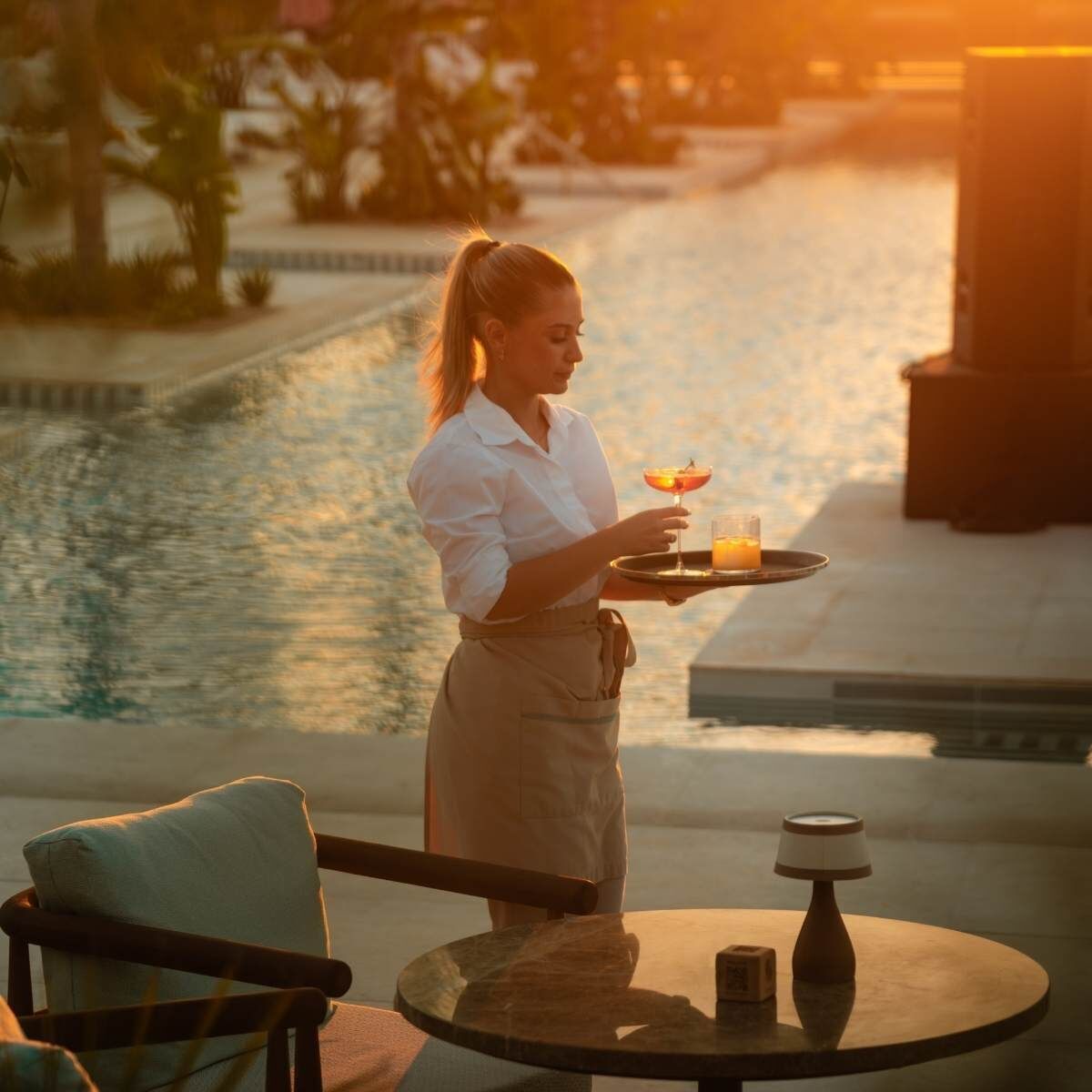 A person holding a tray with a drink and a glass, standing by a pool with a chair and a table nearby.