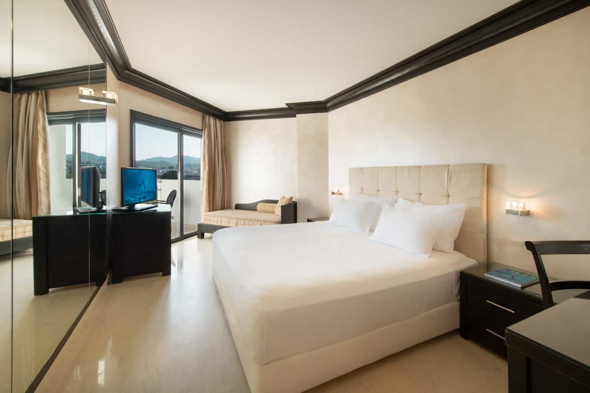 A hotel room with a large bed, a desk with a computer, a chair, a bedside table, and a view through a glass wall.