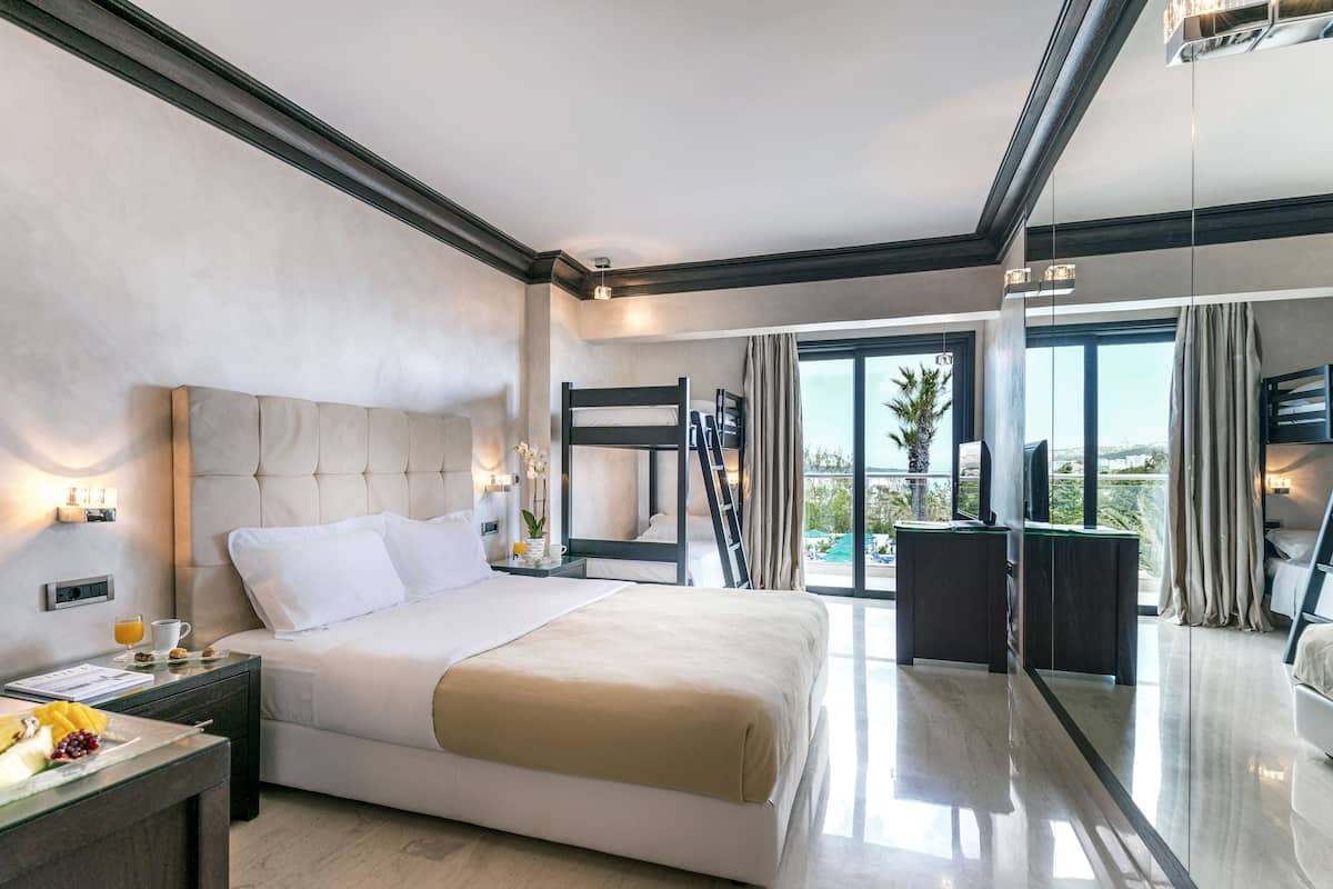 A modern hotel room with a large bed, a bedside table with a lamp, a small table with a cup and a plate of fruit, and a large mirror reflecting the room and a balcony with a pool view.