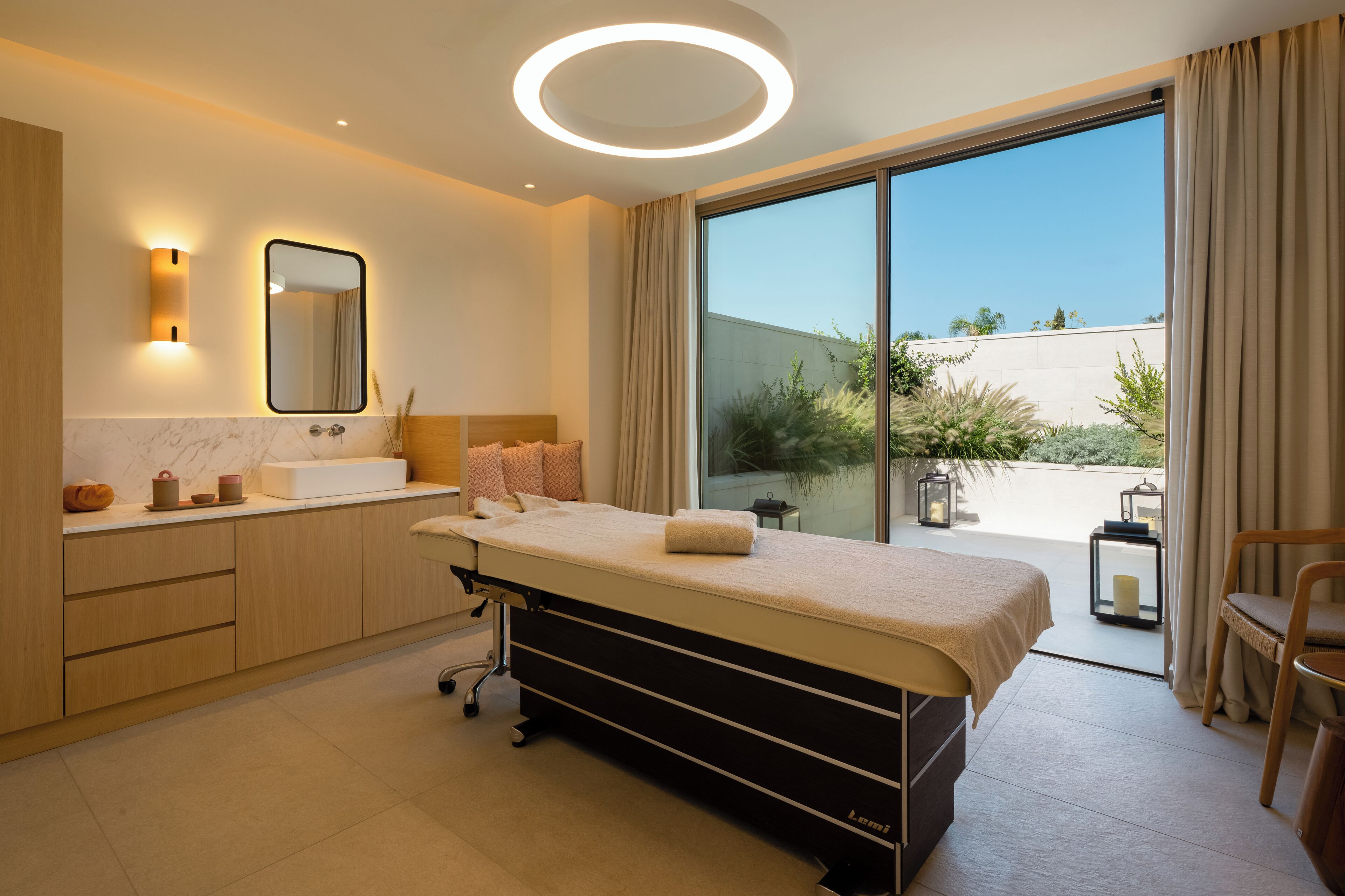 A modern bedroom with a large bed, a bedside table, a mirror, and a view of the outdoors.