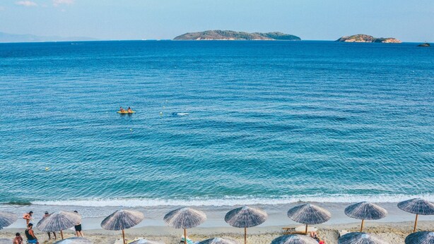 Uma praia com águas azul-claras, uma fileira de guarda-sóis de palha e algumas pessoas na água.