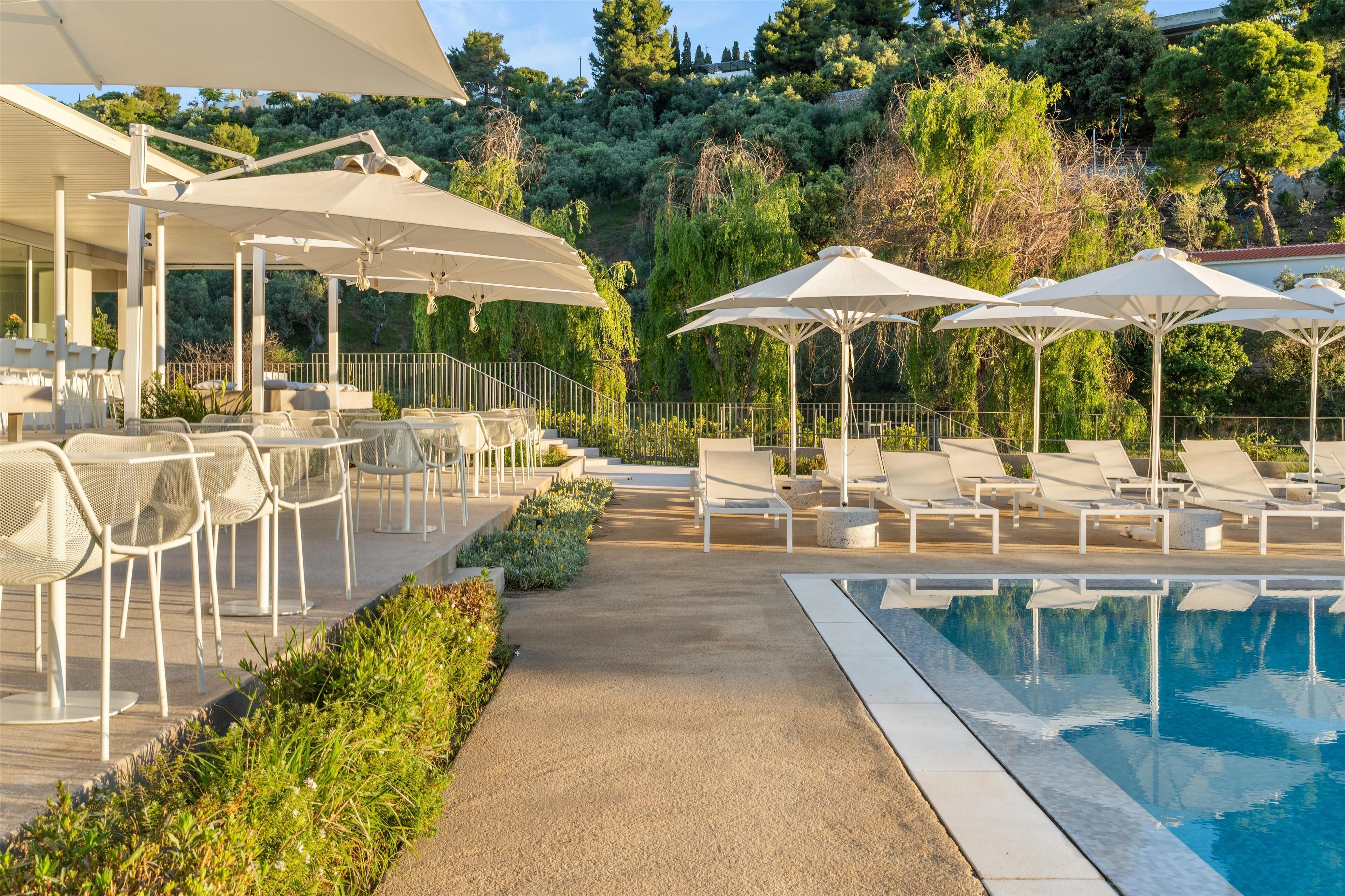 A poolside area with lounge chairs and umbrellas, surrounded by greenery and a building in the background.