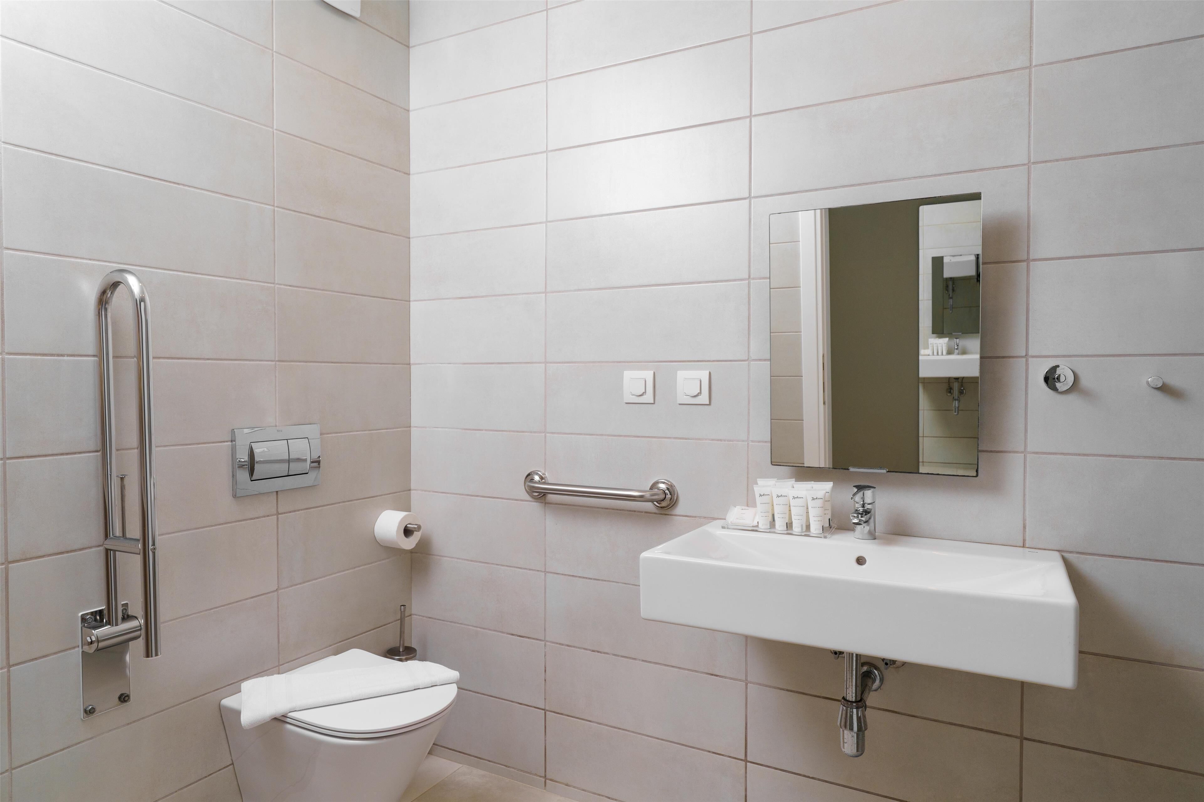 Un baño con inodoro, lavamanos, espejo y barra de apoyo.