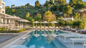 Área de piscina com espreguiçadeiras e guarda-sóis, cercada por vegetação e edifícios.