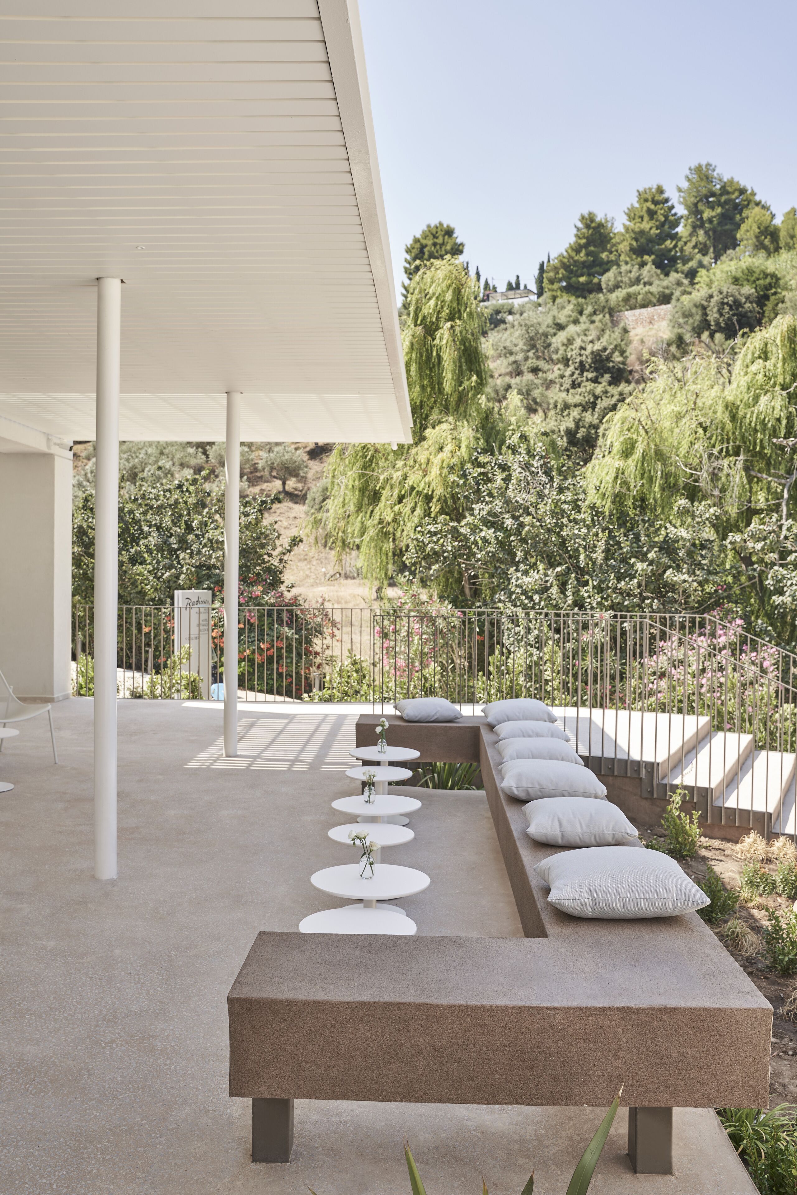 A modern outdoor seating area with a bench and round tables, overlooking a landscaped garden and hillside.
