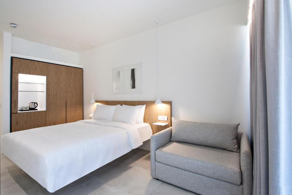 A hotel room with a bed, a sofa, a wooden wardrobe, a nightstand, and a wall-mounted light fixture.