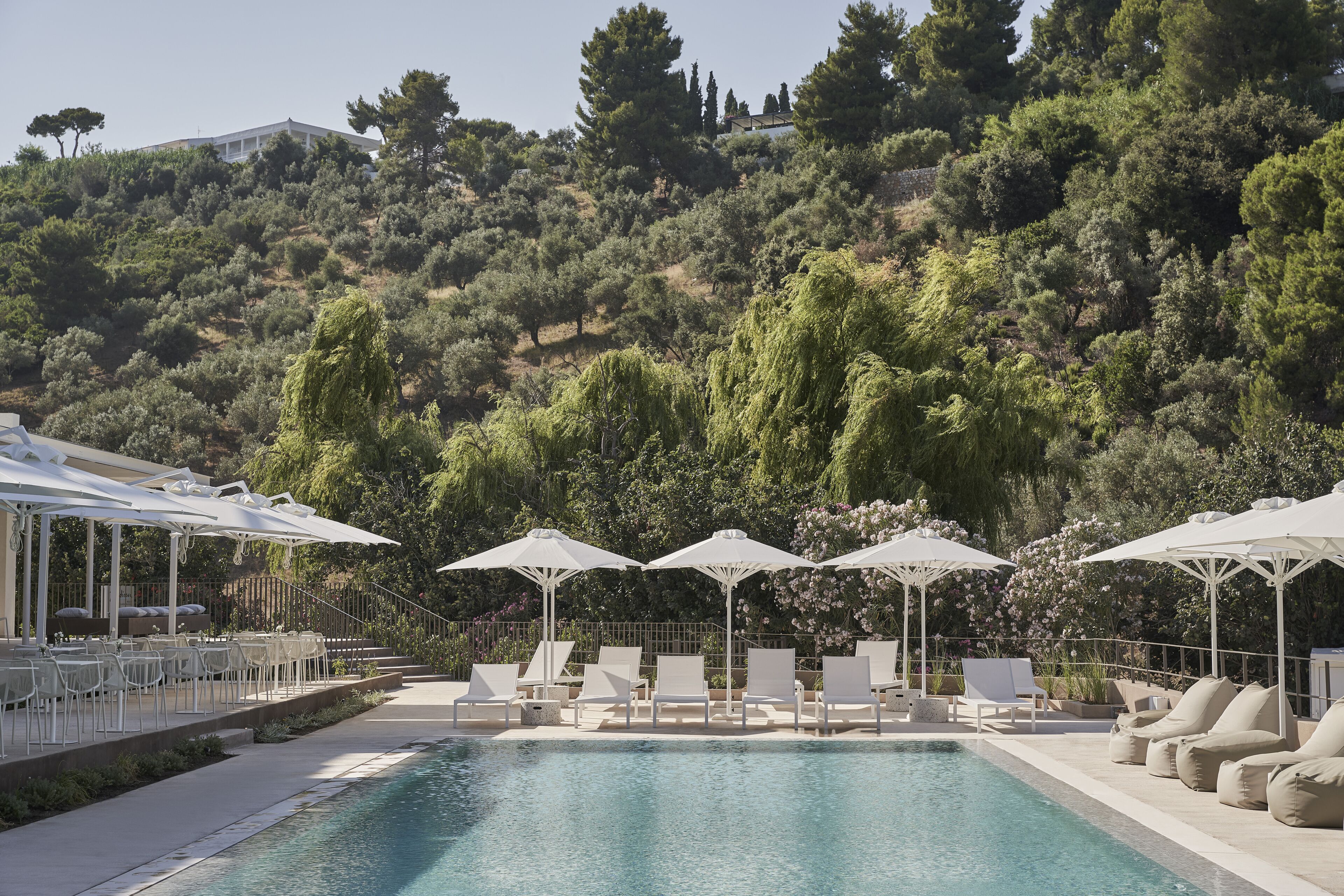 A pool area with lounge chairs and umbrellas, surrounded by a hillside with trees and a building in the background.