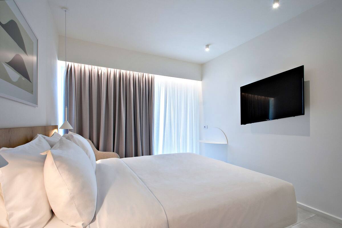 A modern hotel room with a large bed, a flat-screen TV mounted on the wall, and a large window with curtains.