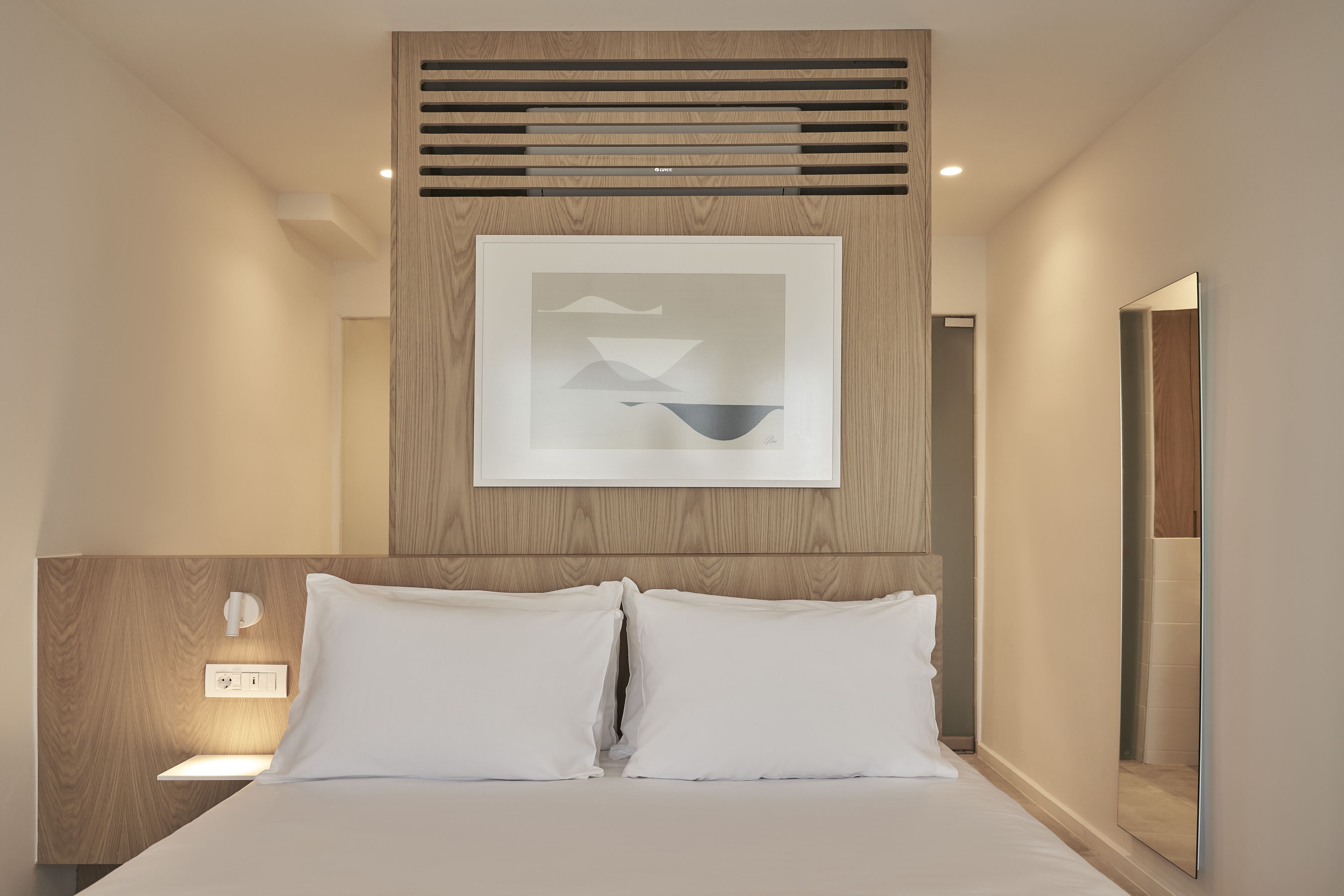 A modern hotel room with a wooden headboard, a framed abstract artwork, a bedside table with a lamp, and a large mirror.