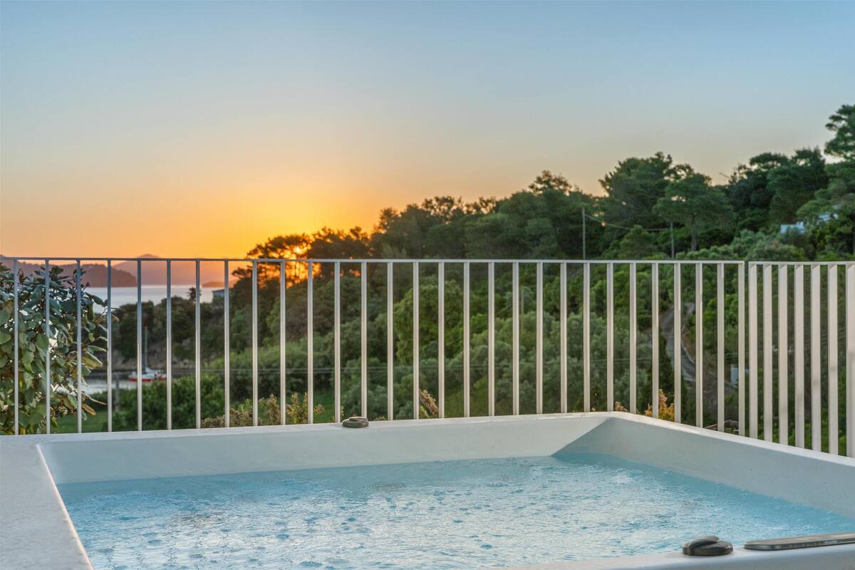 A rectangular hot tub with a white railing, surrounded by greenery and a sunset view.