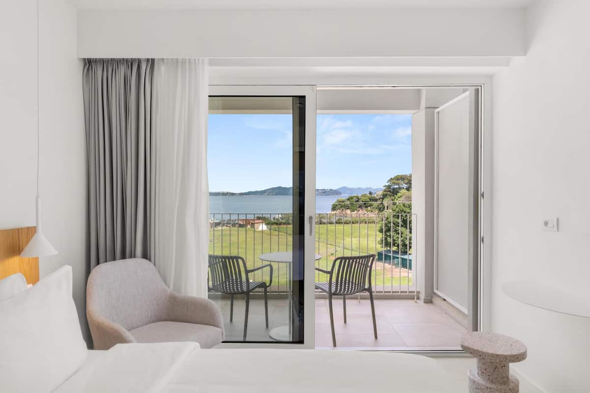A hotel room with a bed, a chair, a small table, and a balcony with a view of the sea and greenery.