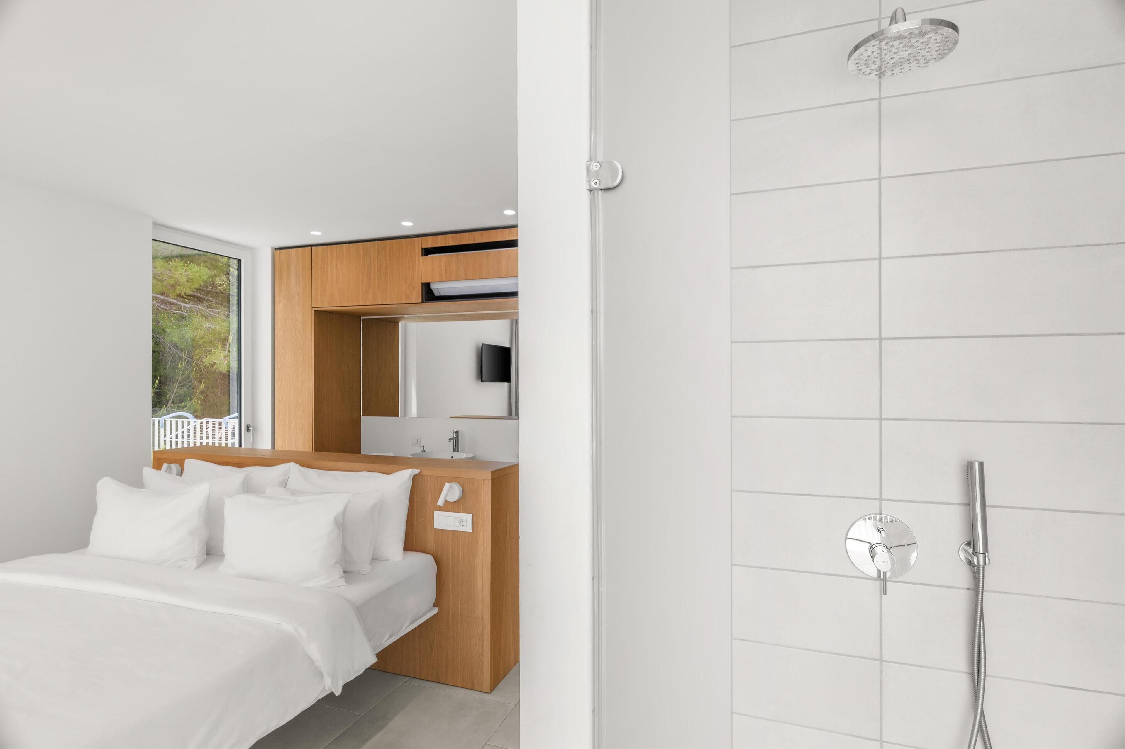 A modern hotel room with a bed, a wooden cabinet, a bathroom with a shower, and a large window.