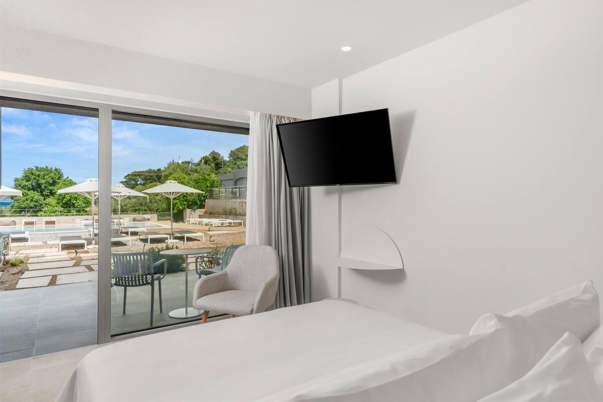 A hotel room with a large window, a bed, a TV, and a view of an outdoor pool and lounge area.