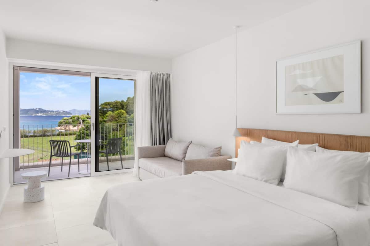 A modern hotel room with a large bed, a sofa, a small table, and a balcony with a view of the sea.