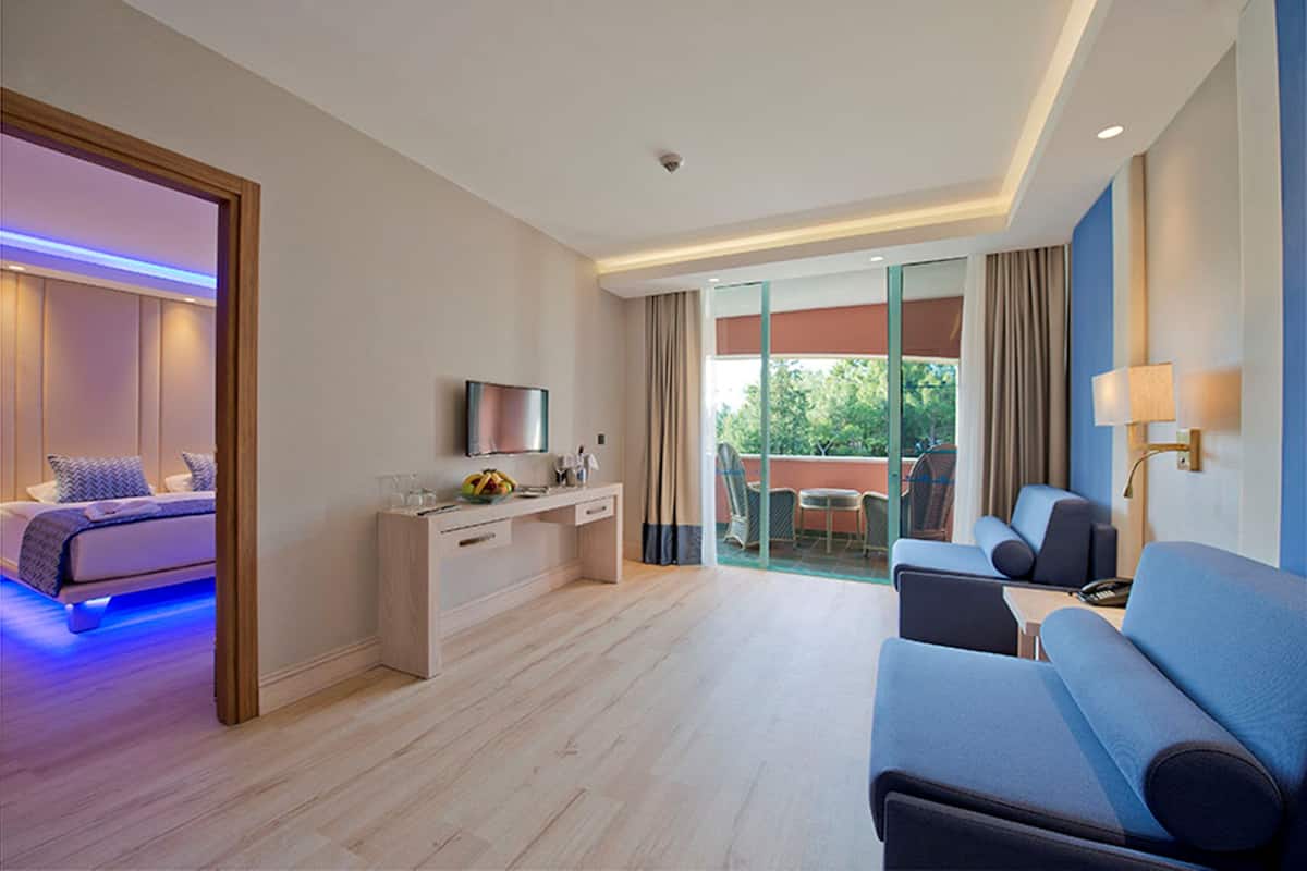 A modern hotel room with a bed, a desk with a flat-screen TV, a sofa, and a balcony with a view of greenery.