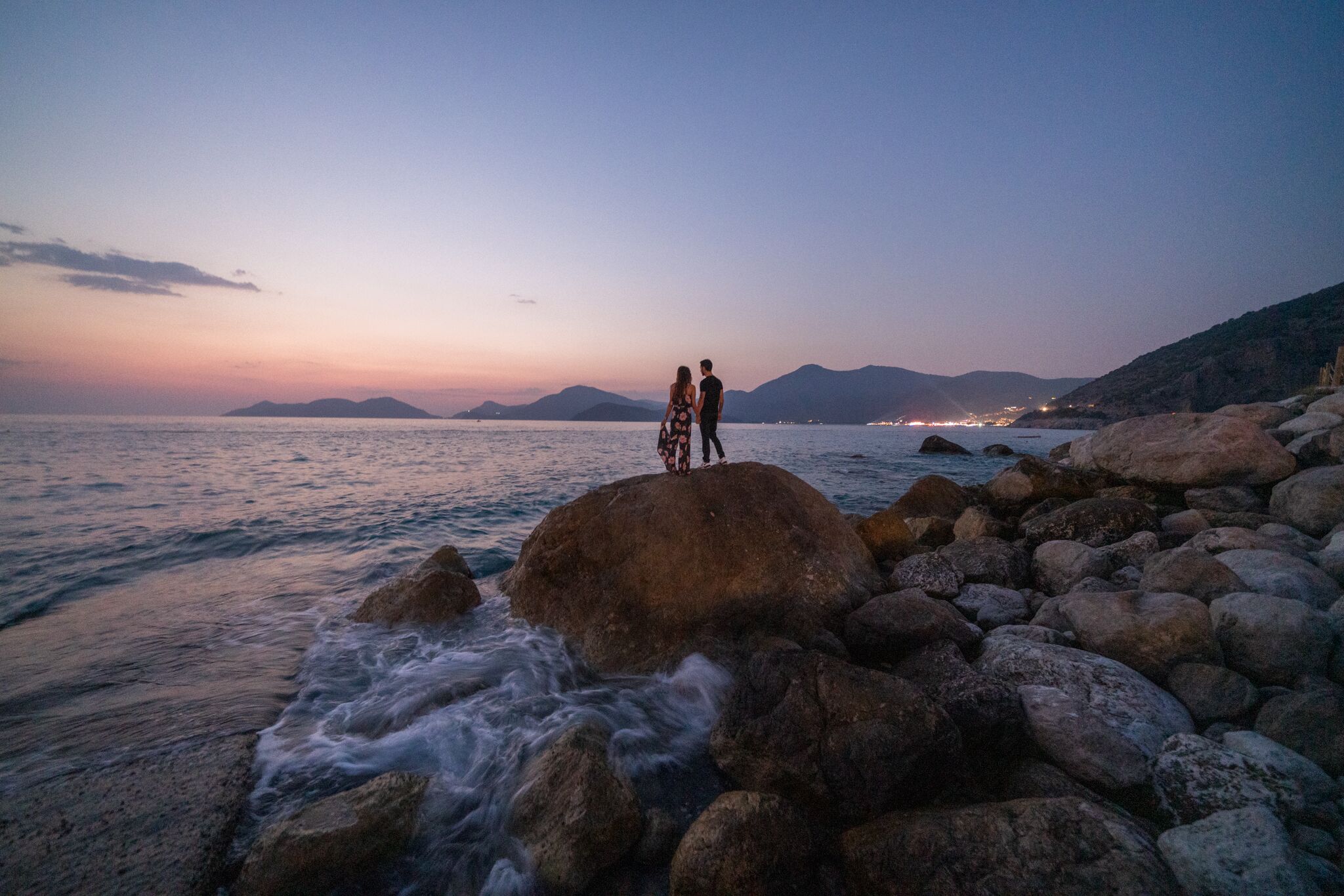 Um casal em cima de um afloramento rochoso, à beira-mar, ao pôr do sol.
