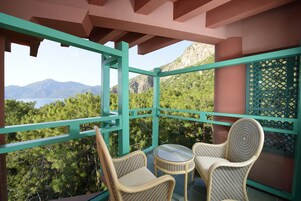 Een balkon met rieten stoelen en een kleine tafel, met uitzicht op een bergachtig landschap en een wateroppervlak.
