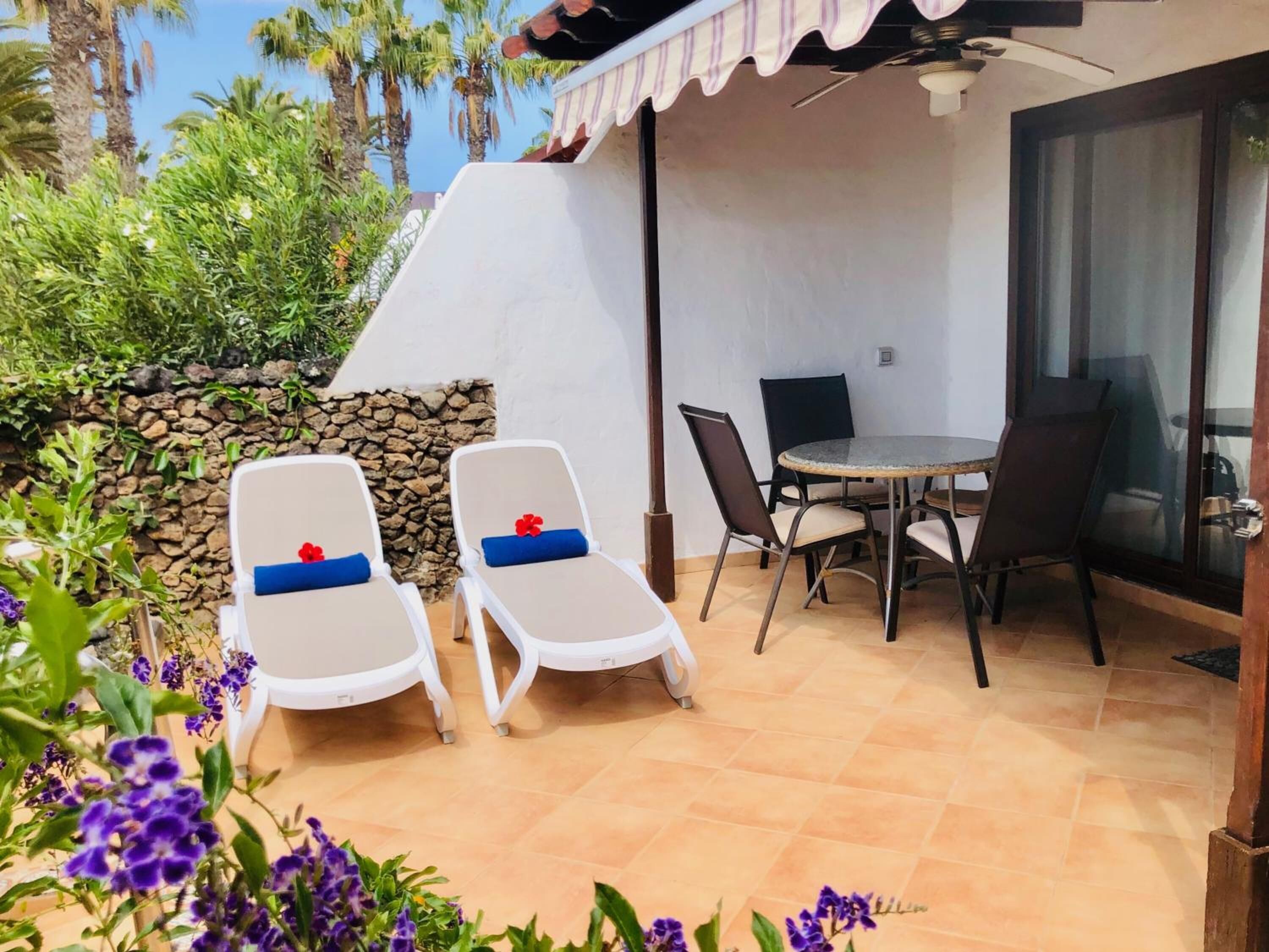Eine Terrasse mit zwei weißen Liegestühlen, einem Tisch mit Stühlen, einer Steinmauer und Blick auf Palmen.