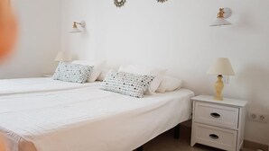 Un dormitorio con dos camas, ropa de cama blanca, almohadas estampadas, lámparas de noche y una mesita de noche con cajones.