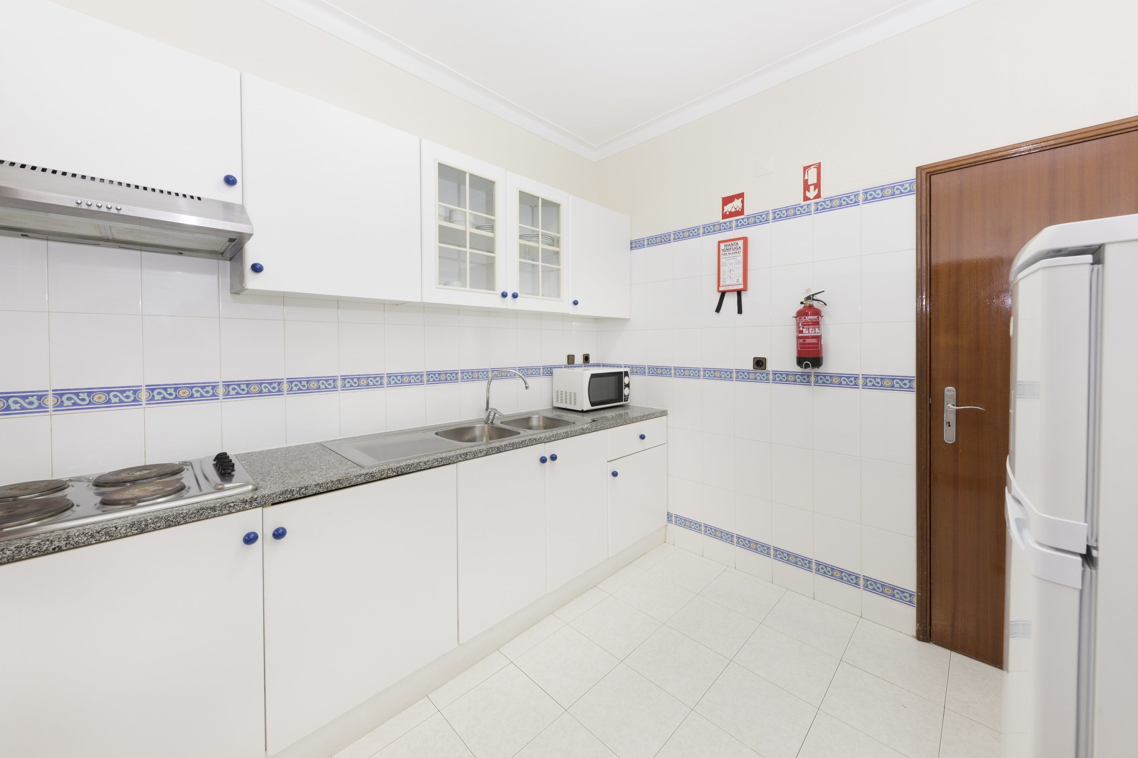 Een keuken met witte kastjes, een magnetron, een fornuis en een spoelbak. Er hangt een brandblusser aan de muur en bevindt zich een deur rechts.