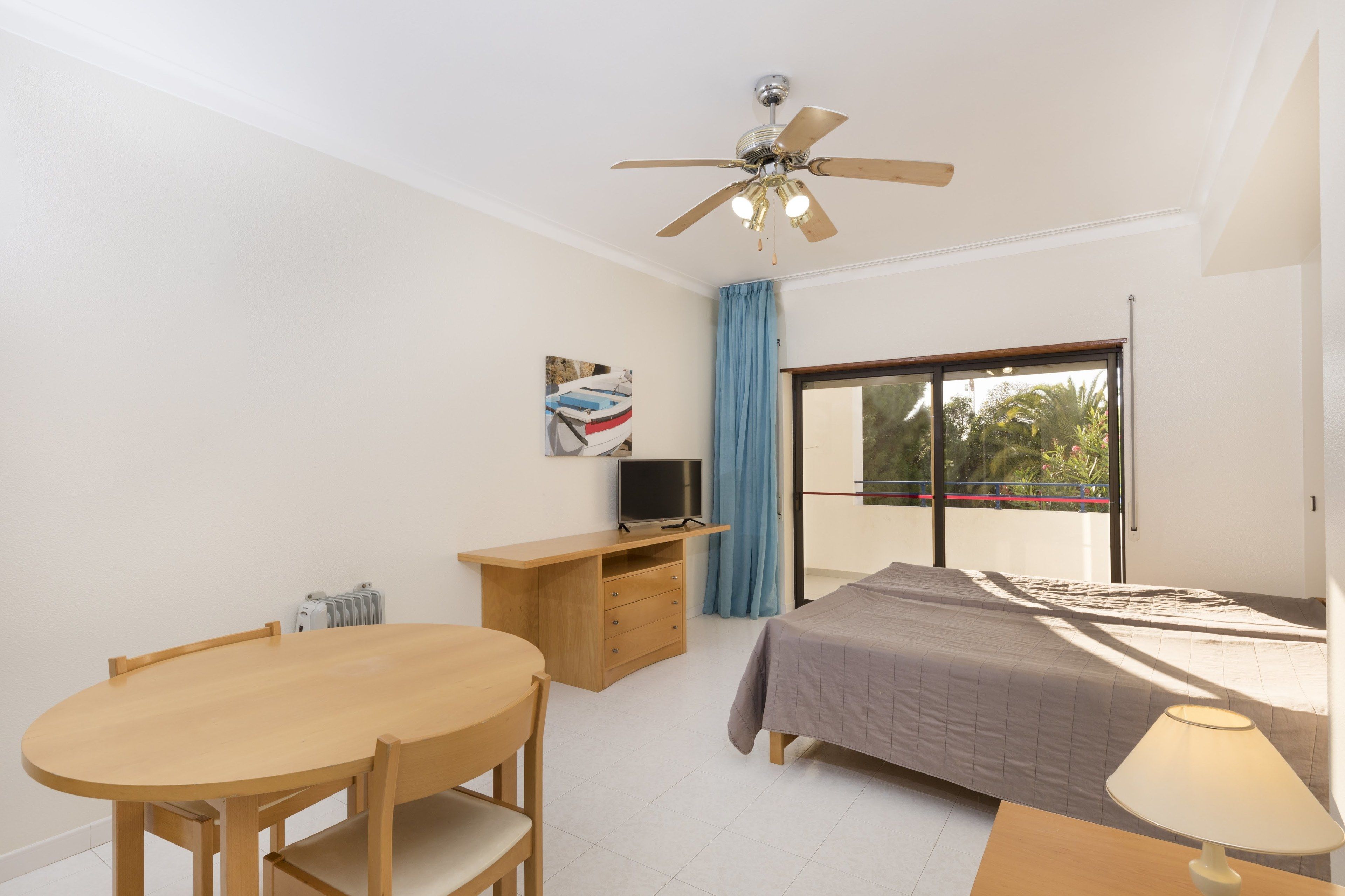 A bedroom with a bed, a desk, a chair, a ceiling fan, a TV, and a window with a view of palm trees.