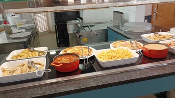 Un mostrador de buffet con diversas opciones de comida, incluyendo pasta, una olla roja de sopa y pizza.