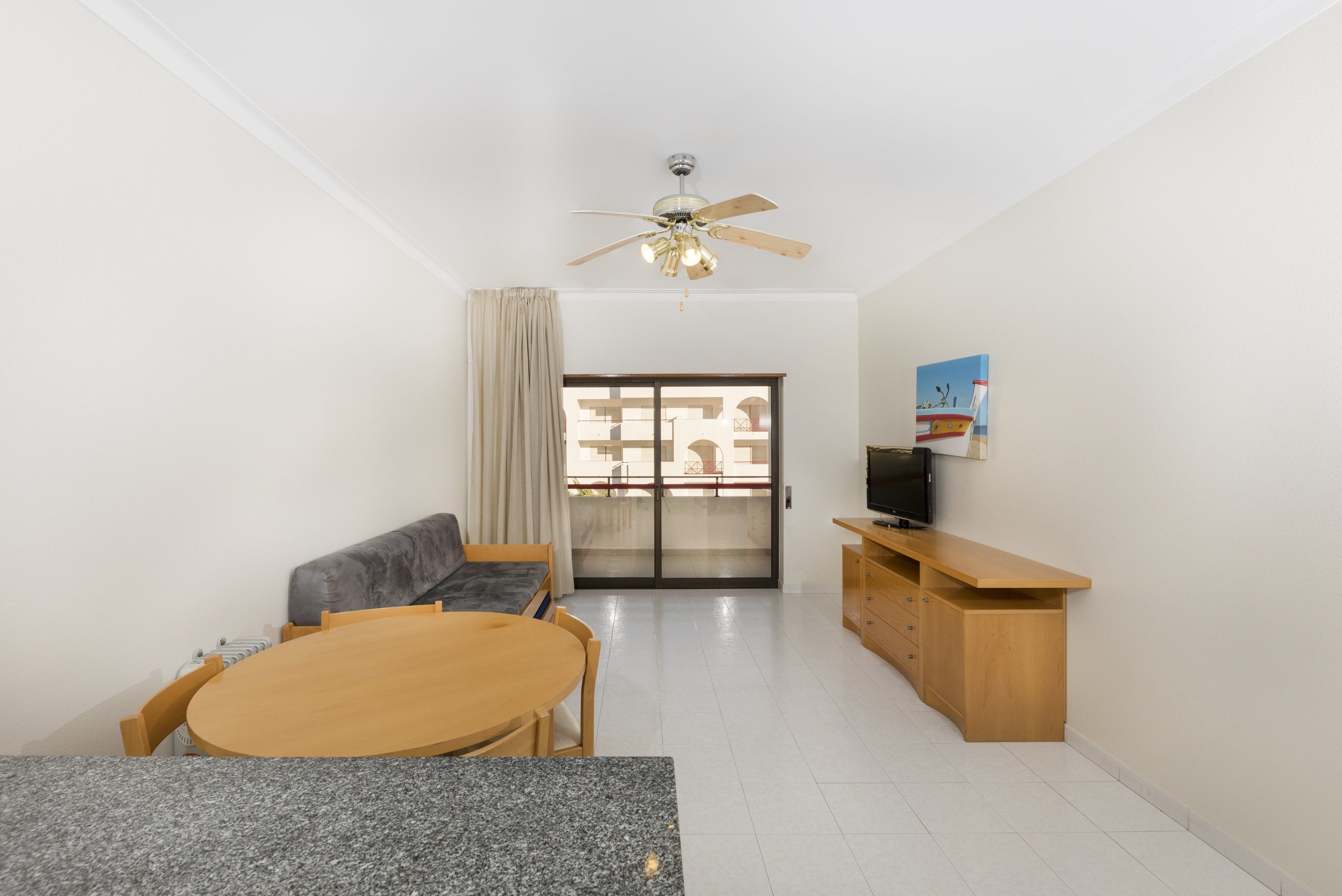 A modern living room with a wooden dining table, a sofa, a ceiling fan, and a TV on a wooden cabinet.