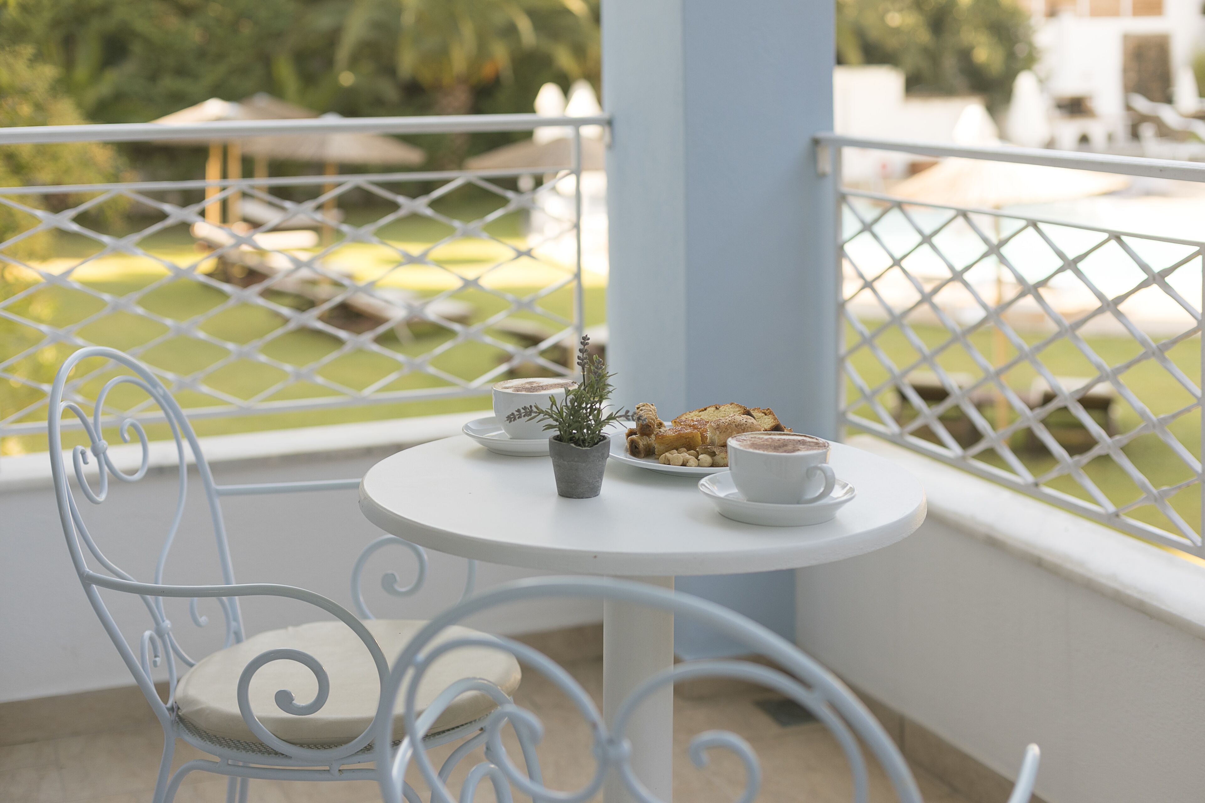 Une chaise et une table en métal blanc, avec une petite plante en pot, une assiette de nourriture et une tasse, sur un balcon.