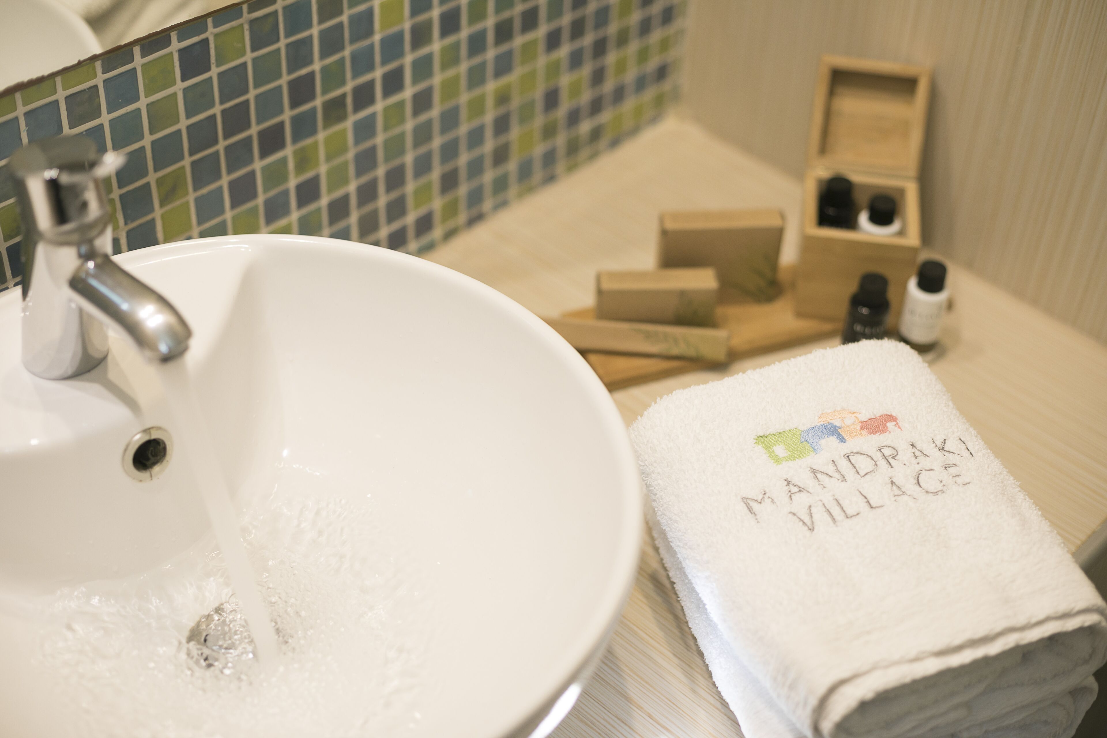 Ein Badezimmer mit einem weißen Waschbecken, einem Handtuch mit dem Schriftzug 'MANDRAKI VILLAGE' und einer Holzkiste mit Pflegeprodukten.